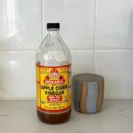 A bottle of Bragg organic apple cider vinegar sits next to a gray and brown ceramic mug on the marble kitchen counter.