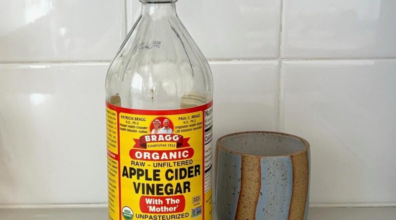 A bottle of Bragg organic apple cider vinegar sits next to a gray and brown ceramic mug on the marble kitchen counter.