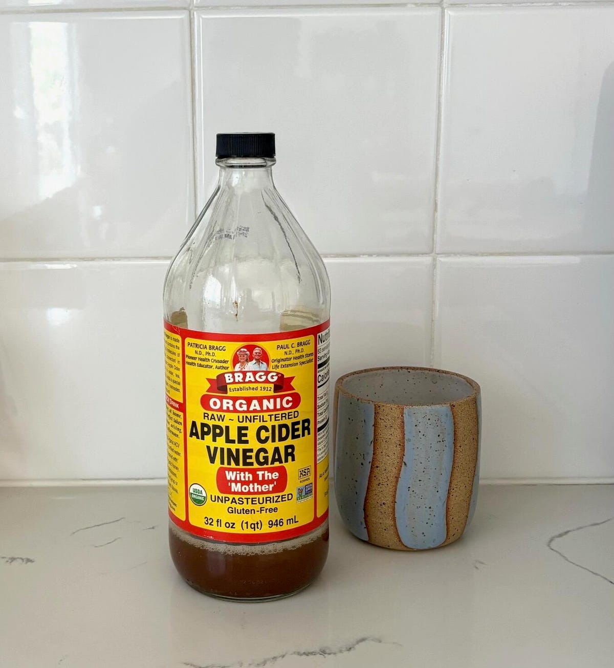 A bottle of Bragg organic apple cider vinegar sits next to a gray and brown ceramic mug on the marble kitchen counter.